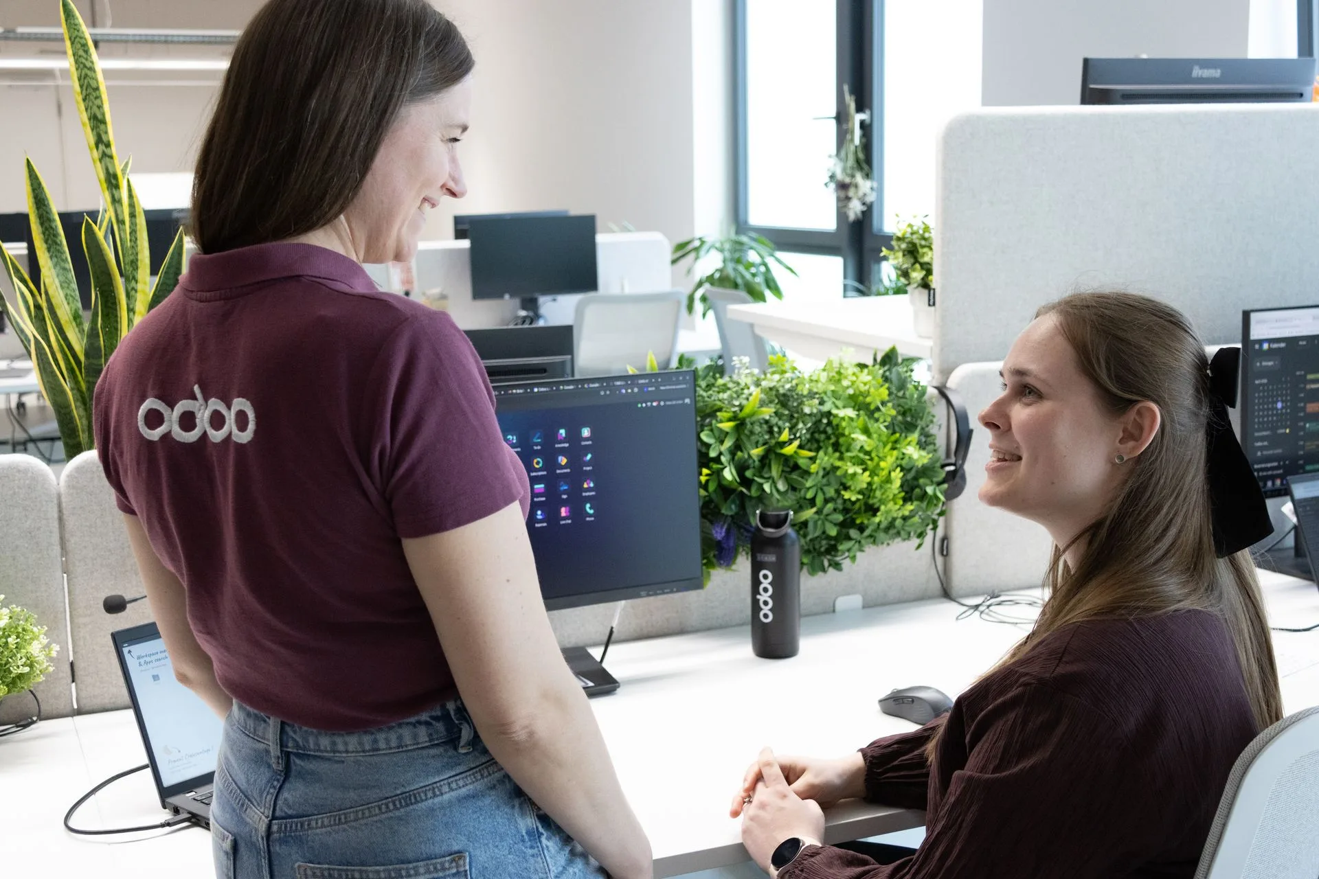 Співробітниця Odoo в фіолетовому поло усміхнено спілкується з колегою за робочим столом у сучасному офісі